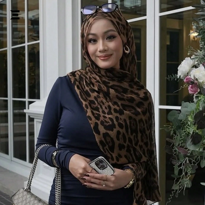 Woman wearing a leopard print hijab and navy blue top, holding a phone and a handbag, standing in front of a building with flowers.