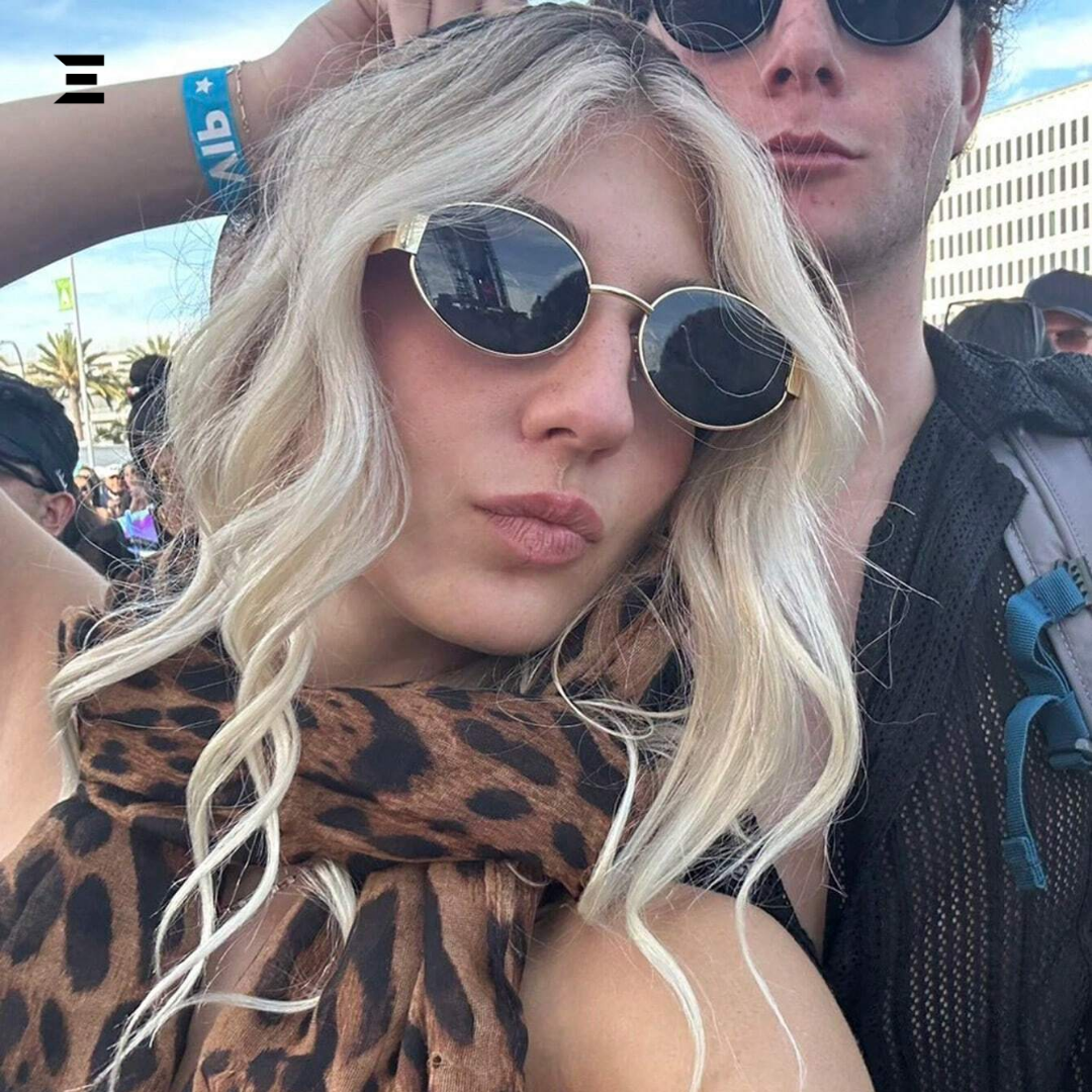 Person with blonde hair and sunglasses wearing a leopard print scarf, with a blurred background of people and buildings.