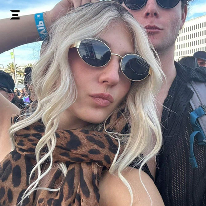 Person with blonde hair and sunglasses wearing a leopard print scarf, with a blurred background of people and buildings.